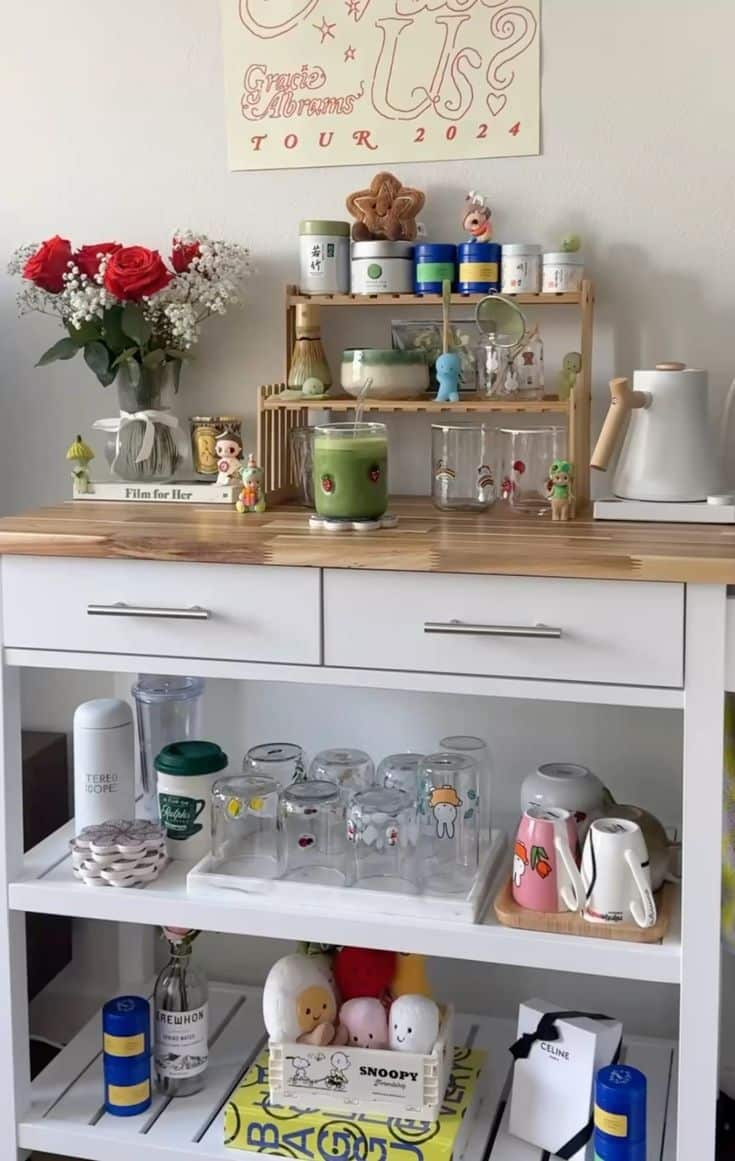 White console-style matcha bar with layered shelves holding mugs, plush toys, glassware, and a bamboo matcha station, styled with red roses and cute figurines.