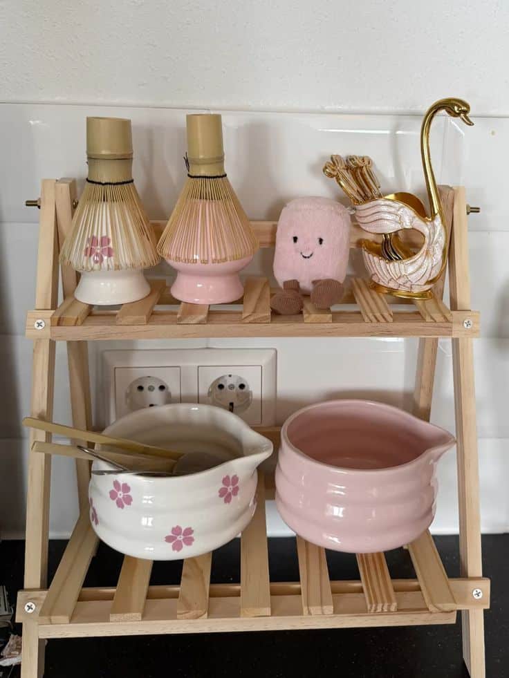 Pastel matcha bar setup with pink and white bowls, bamboo whisks, and cherry blossom patterns on a wood shelf.