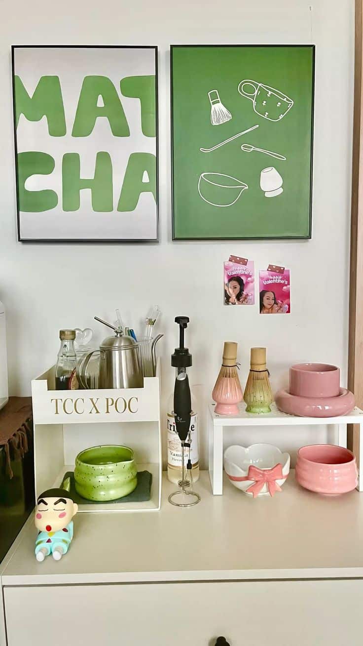 Matcha station with framed green art, electric frother, matcha tools, and pink ceramic cups on white shelves.