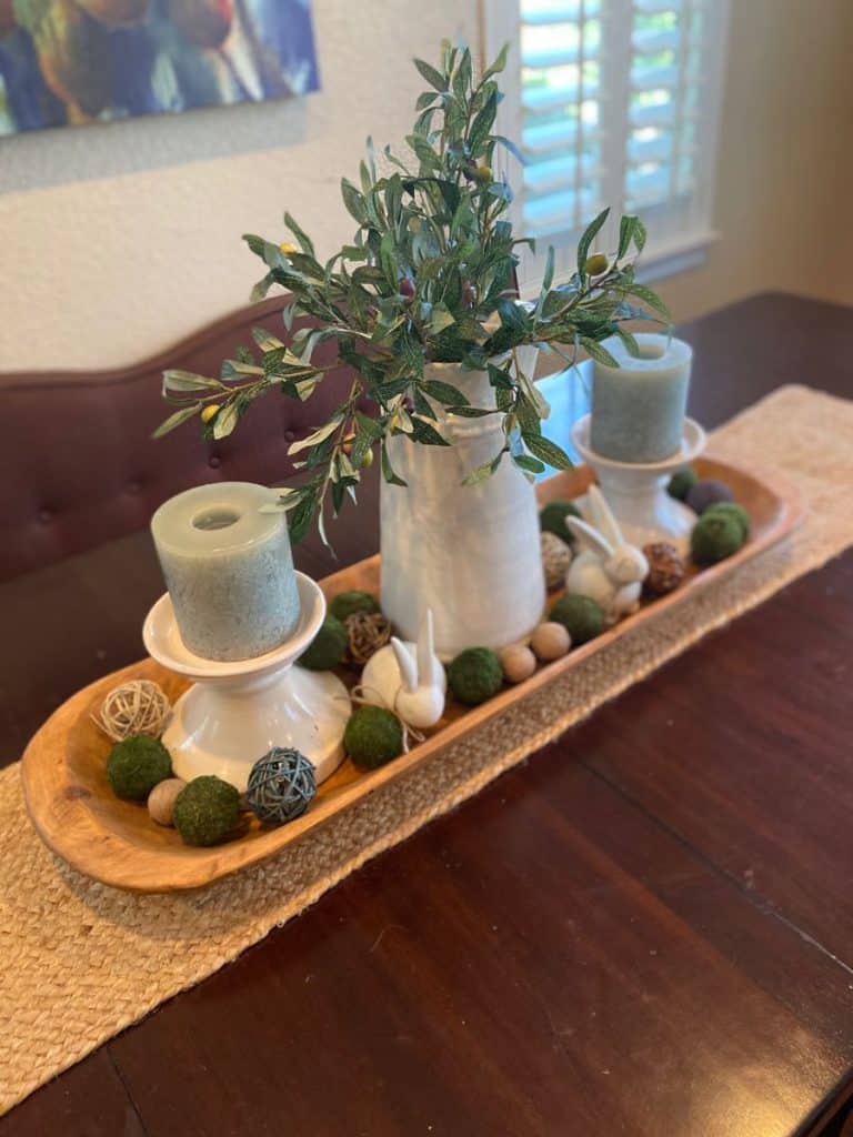 Spring Easter dough bowl centerpiece with white ceramic bunnies, olive branches in a jug, and dusty blue candles.