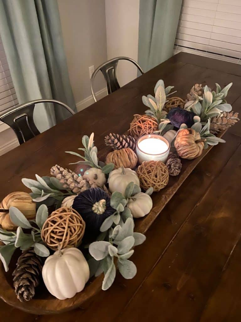 Dark wooden dough bowl centerpiece with velvet pumpkins, rattan balls, candle, pine cones, and lambs ear.
