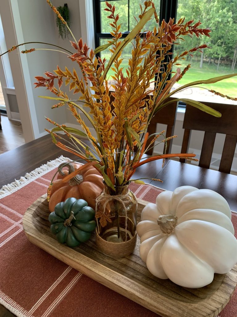 Simple autumn dough bowl centerpiece with a glass vase of tall orange and yellow grasses and three faux pumpkins.