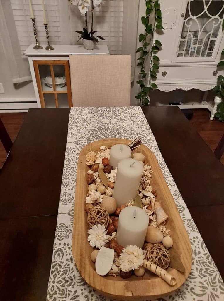 Oval dough bowl centerpiece with three cream pillar candles and natural, scented potpourri with wooden accents.