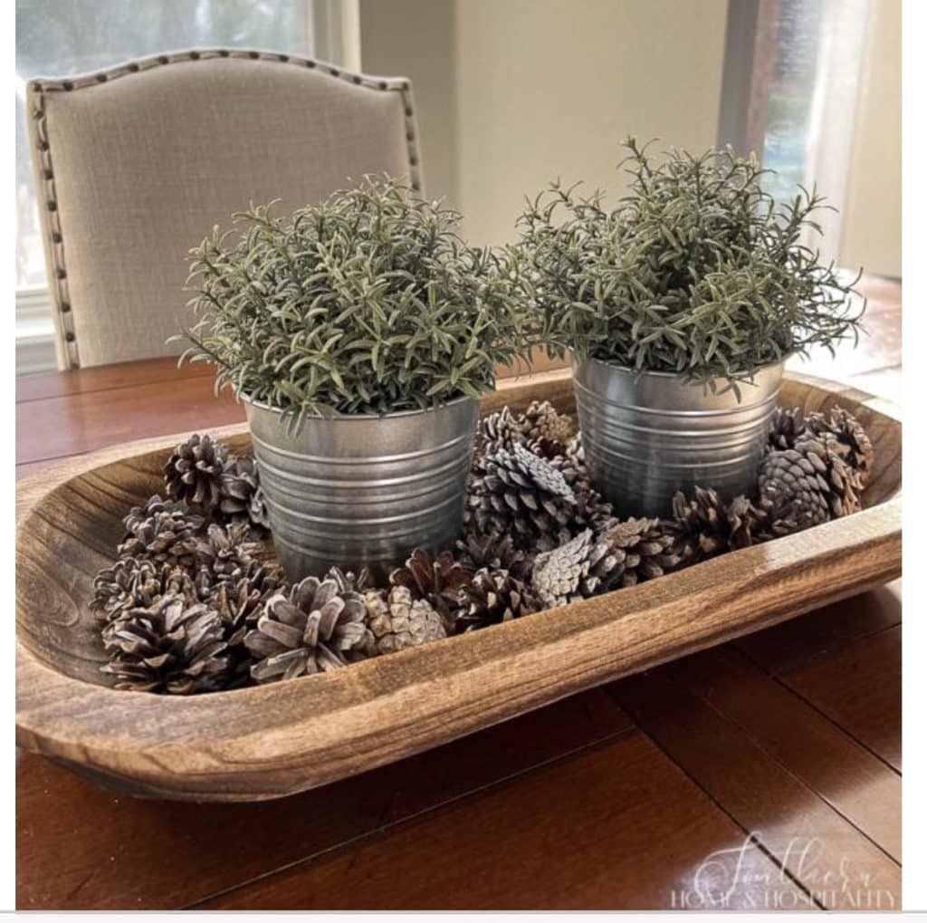 Long wooden dough bowl centerpiece filled with frosted pine cones and two galvanized metal pots of faux rosemary.