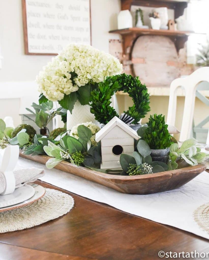 Spring dough bowl centerpiece with white birdhouse, white hydrangea bouquet, boxwood wreath, and trailing greenery.
