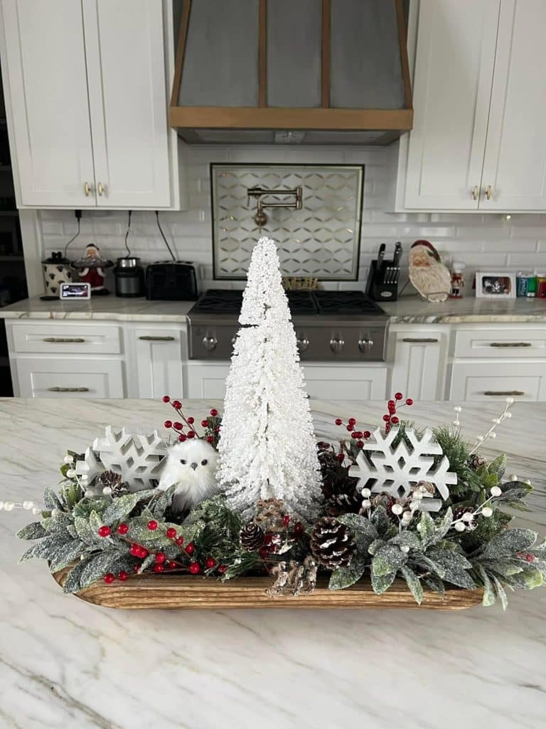 White Christmas dough bowl centerpiece with flocked tree, white owl, snowflakes, and red berries on a marble counter.