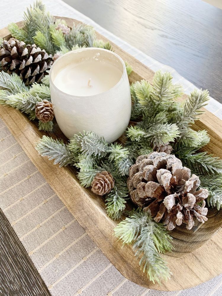 Close-up of a rustic wooden dough bowl centerpiece with a white candle, flocked pine sprigs, and frosted pine cones.