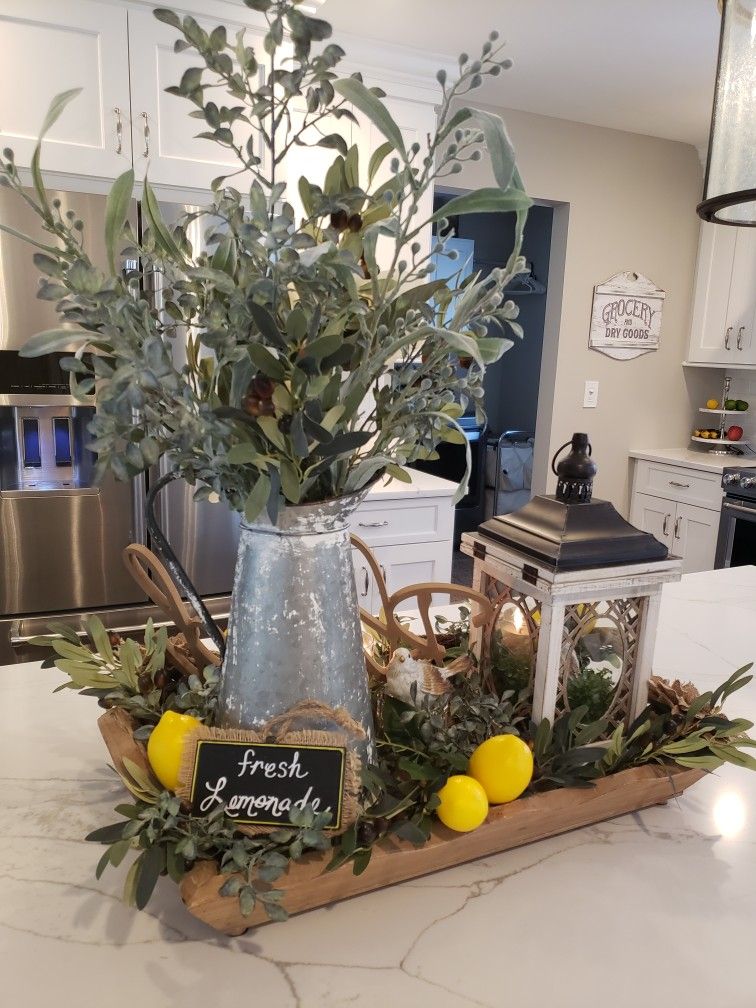 Summer dough bowl centerpiece with a metal lantern, pitcher of greenery, and bright faux lemons on a kitchen island.