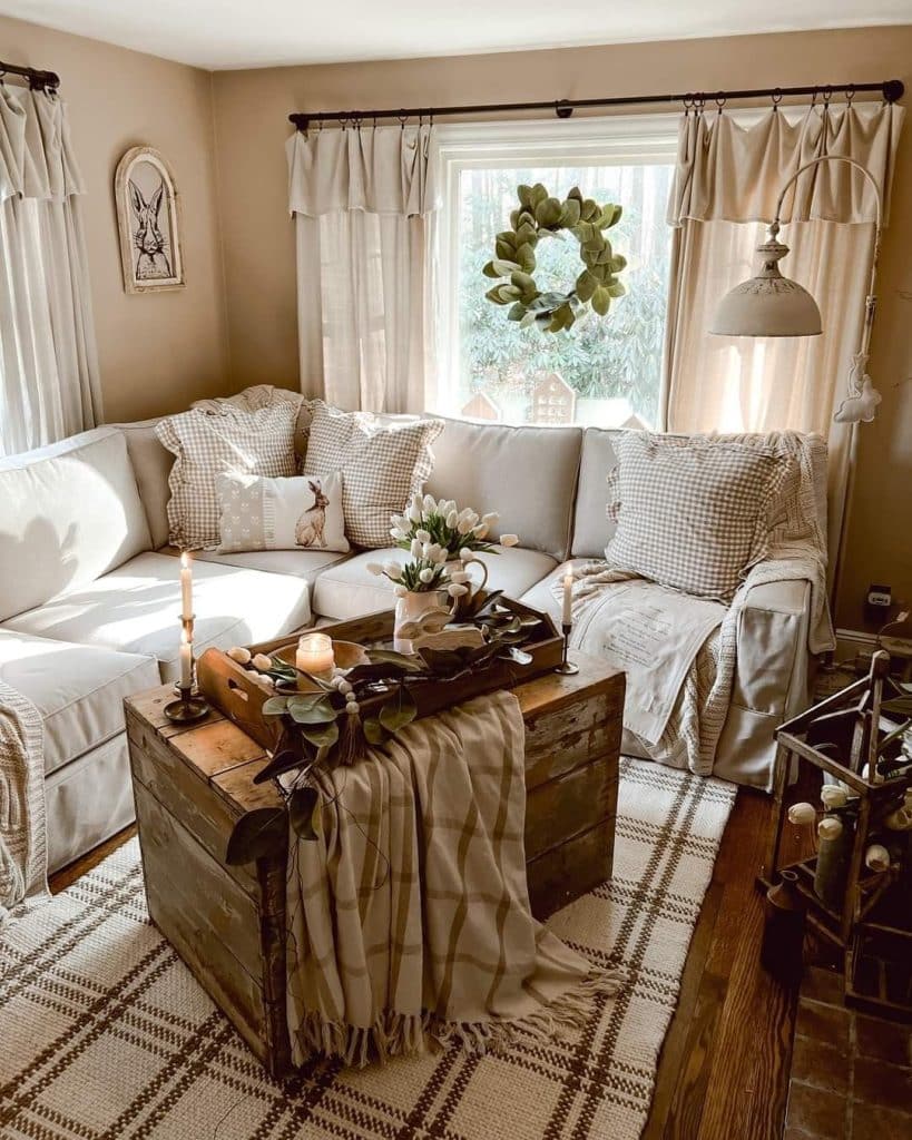 Neutral farmhouse living room with sectional, gingham/check cushions, dark wooden trunk coffee table, and spring wreath in the window.