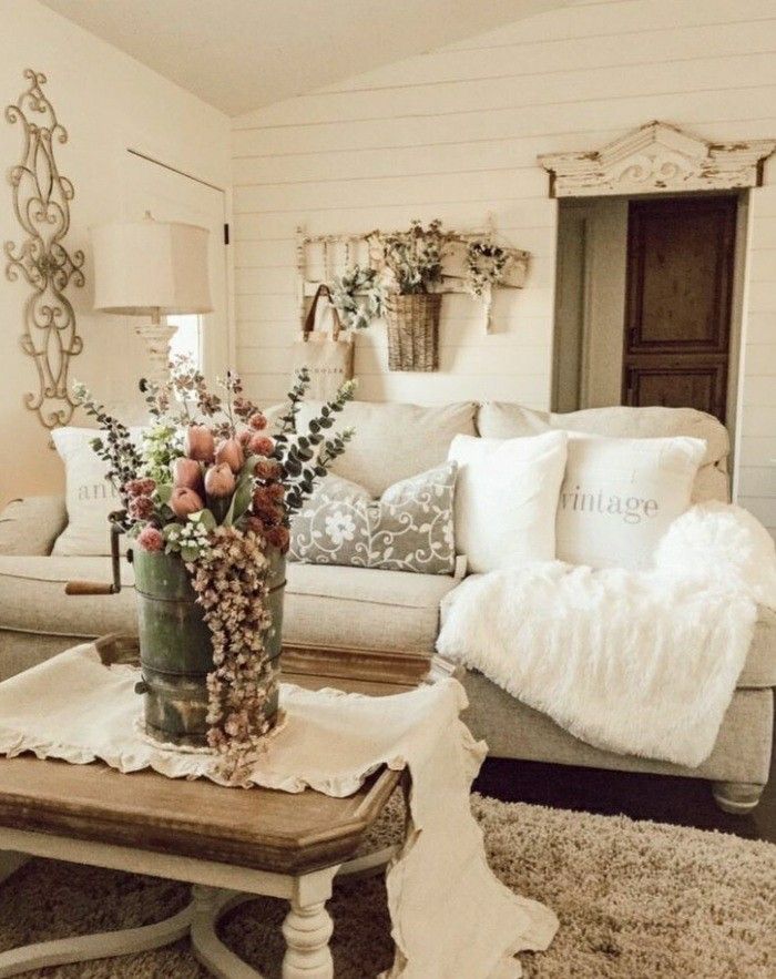 Cozy, neutral farmhouse living room with fuzzy throw, floral cushions, and antique metal vase centerpiece on a wooden coffee table.