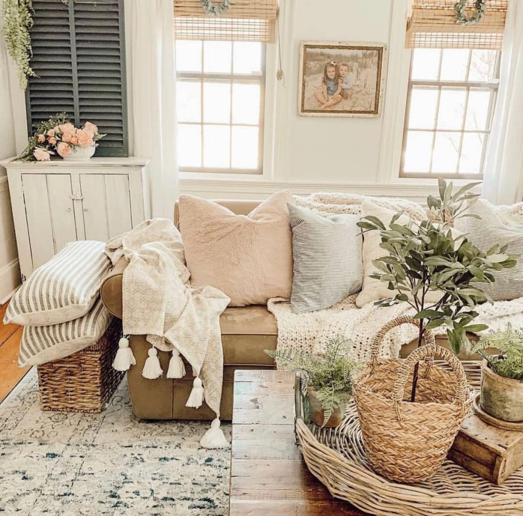 Cozy farmhouse corner with layered pillows, knit throws with tassels, wicker storage basket, and natural plants on a wooden tray.