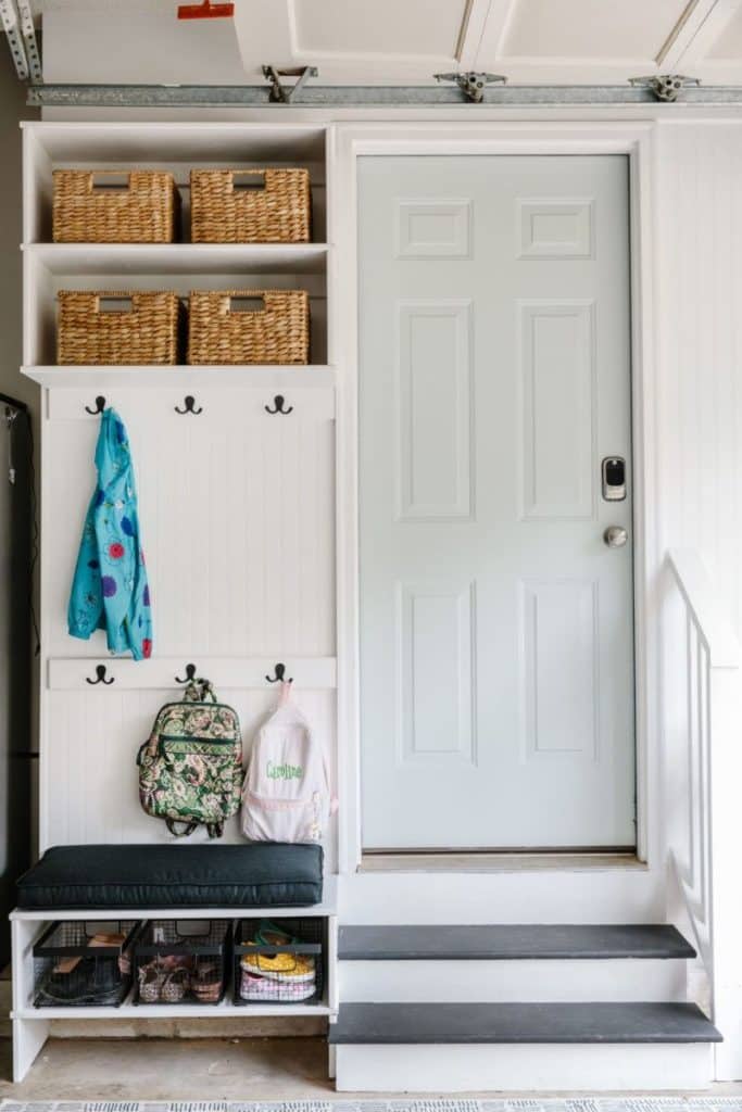 Bright white garage entryway featuring a built-in mud room unit with a dark cushion bench, woven baskets on top shelves, and hooks for backpacks.