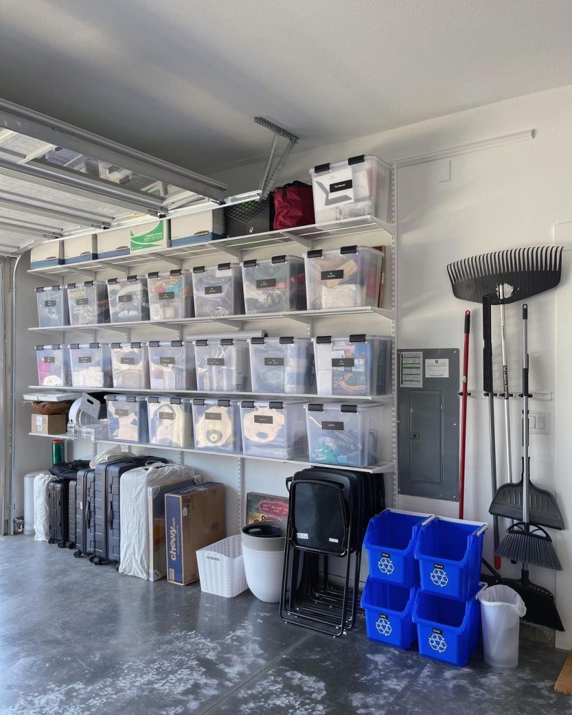 Large garage wall with white track shelving supporting many rows of clear plastic storage containers, with a broom and rake hanging beside.