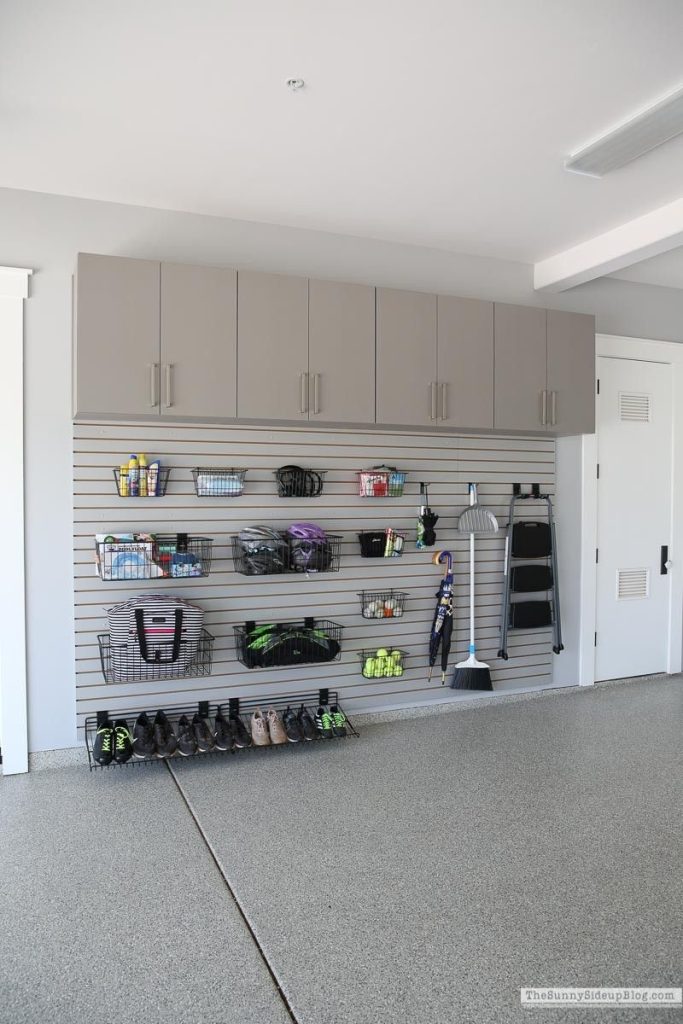 Light gray slat wall system with black wire baskets and hooks, featuring a top row of matching gray storage cabinets.