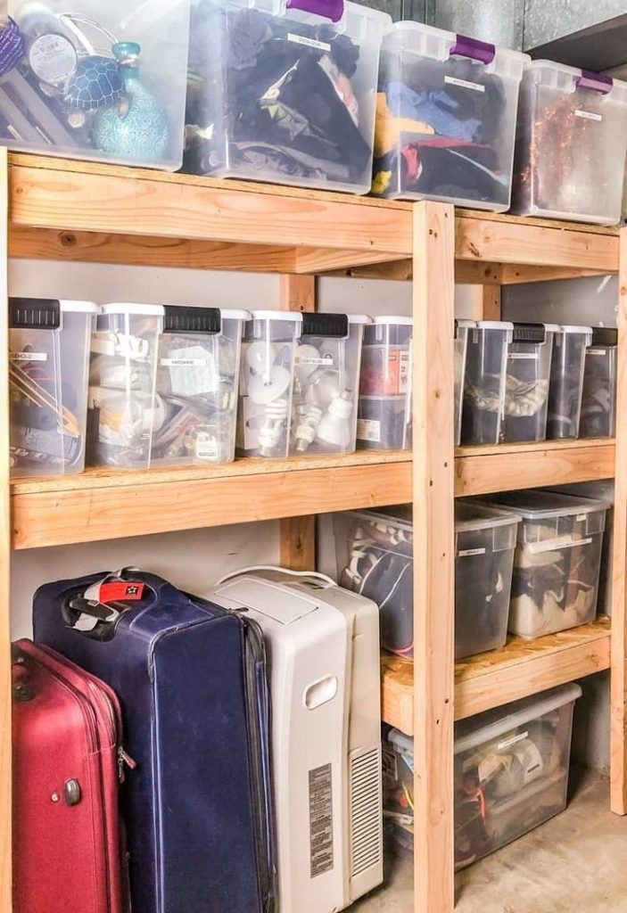 Wooden garage shelving unit with stacked clear plastic storage bins containing out-of-season clothes, luggage, and a portable air conditioner.