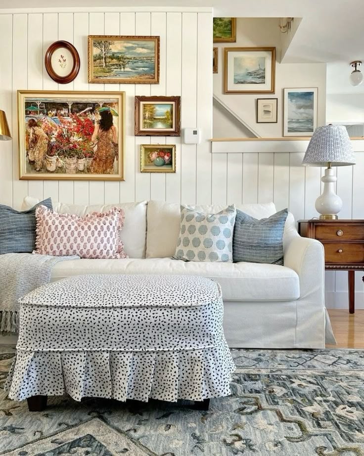 An eclectic gallery of gold-framed art on a white wood-paneled wall above a white sofa.