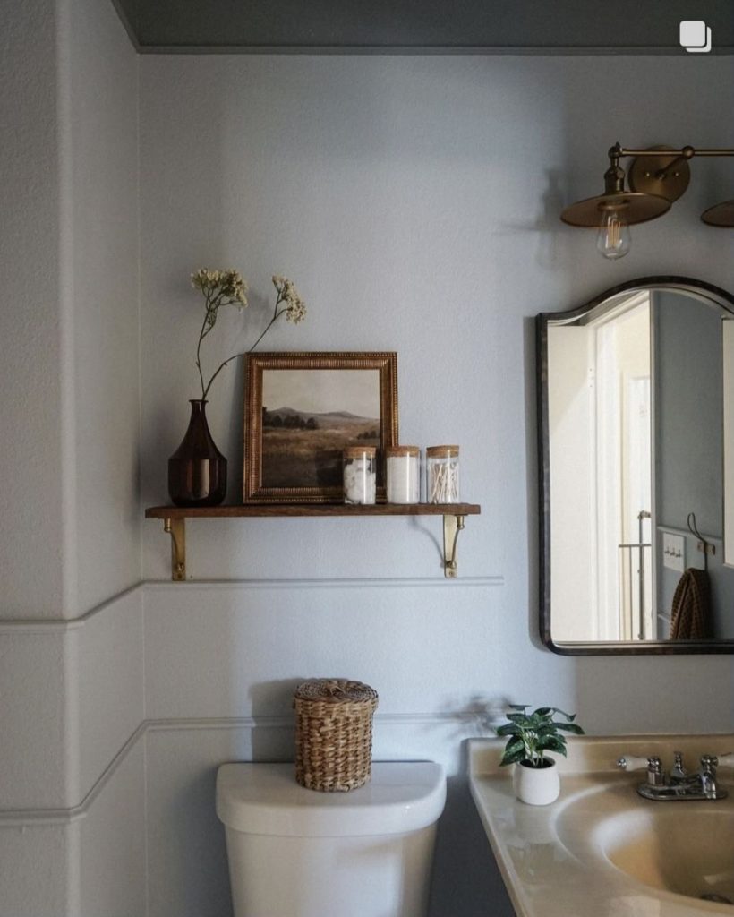 A single dark wood shelf with brass brackets holding a gold-framed landscape painting, glass storage jars, and a tall amber bud vase.