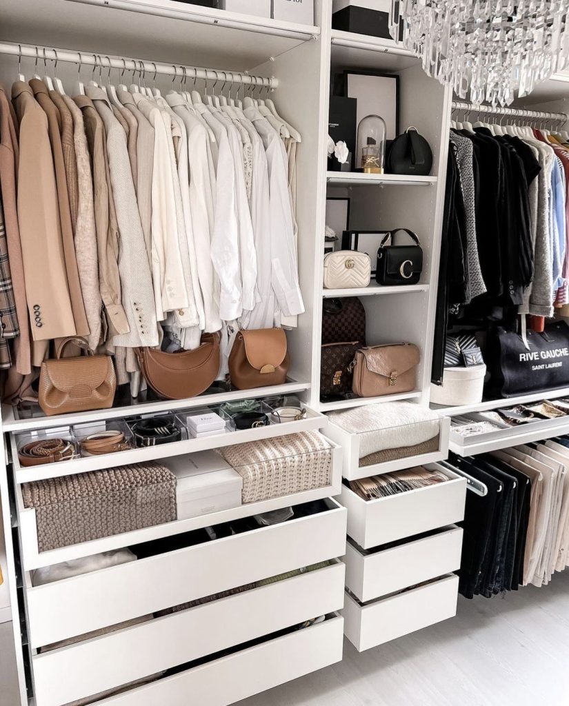 Luxurious walk-in closet with white drawers, wood trim, crystal chandelier, neutral clothing, and designer handbags displayed on shelves.