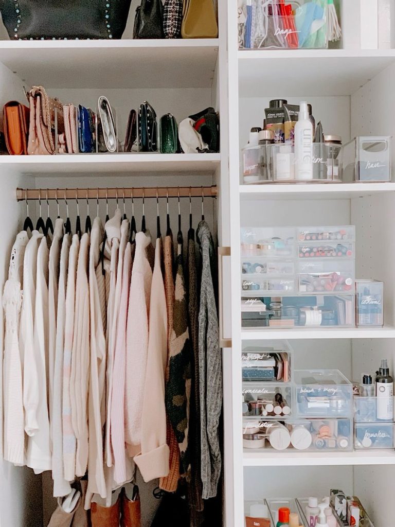 White closet shelving unit with clear acrylic organizers separating makeup, beauty products, and small accessories, next to a rod of neutral clothing.