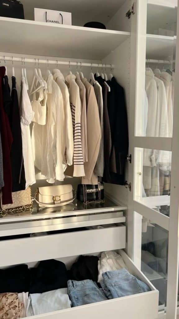 Modern white wardrobe with glass doors, white hangers, neutral clothing, and luxury handbags displayed on a mirrored shelf above drawers.