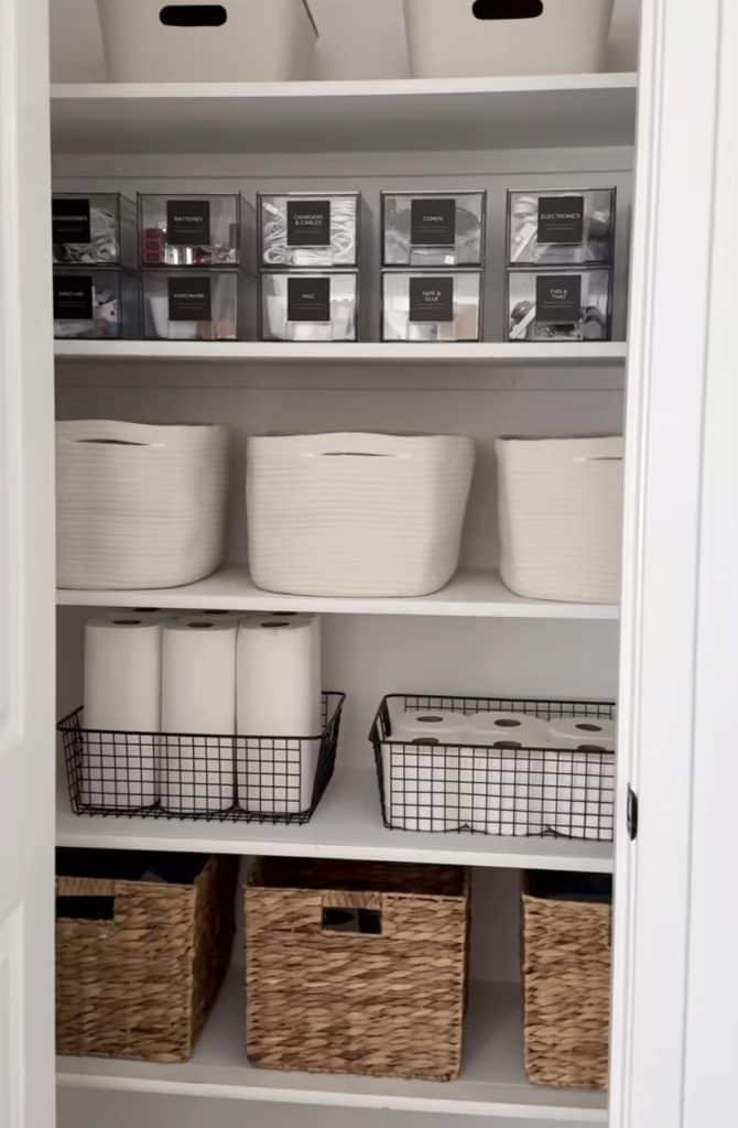 Organized utility closet with shelves of labelled clear bins for hardware and electronics, white rope baskets, and large woven storage bins.