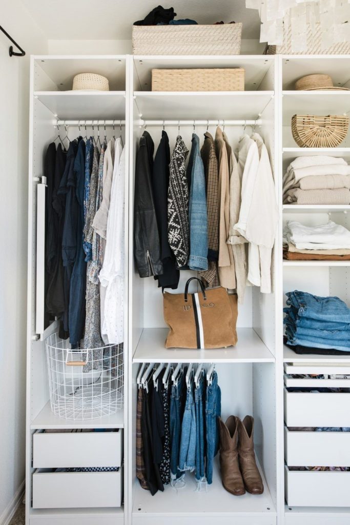 White modular closet system with hanging space, drawers, shelves for folded jeans and sweaters, and open shelving displaying handbags and boots.