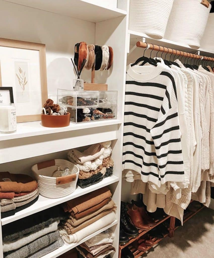 White closet shelf styled with folded knitwear, a small framed print, a diffuser, and a clear box containing hair accessories and headbands on a stand.