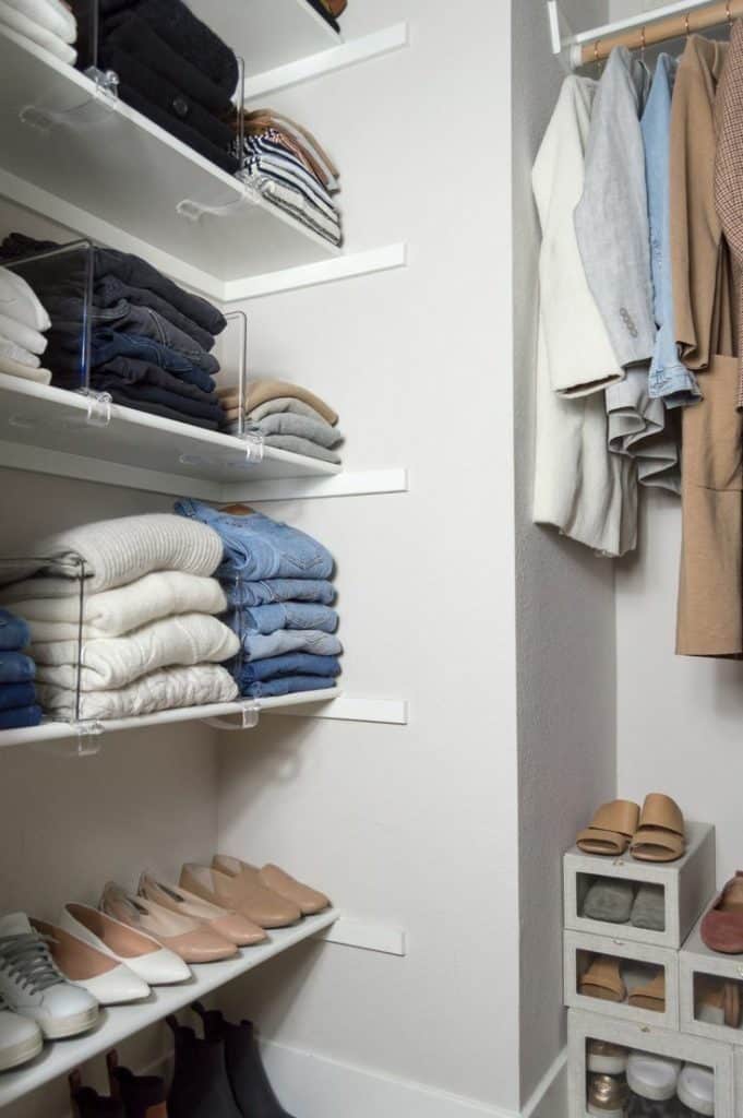 White closet shelving corner with folded sweaters and jeans separated by acrylic dividers, and neat rows of nude ballet flats and loafers below.