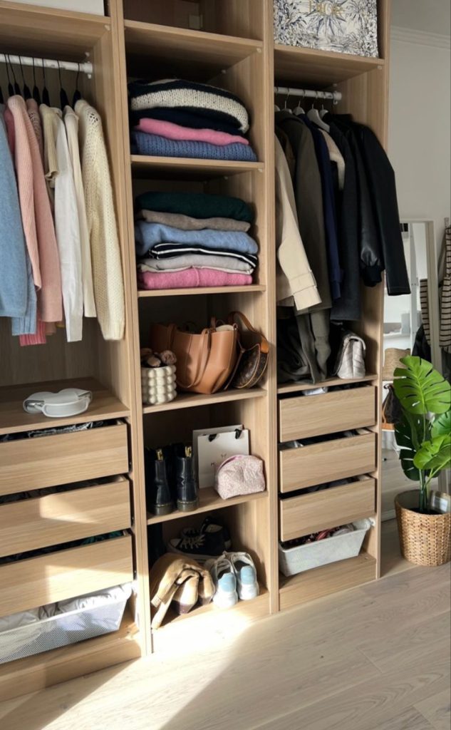 Light wood wardrobe system featuring hanging clothes, stacked sweaters, drawers, and open shelving displaying handbags and boots.