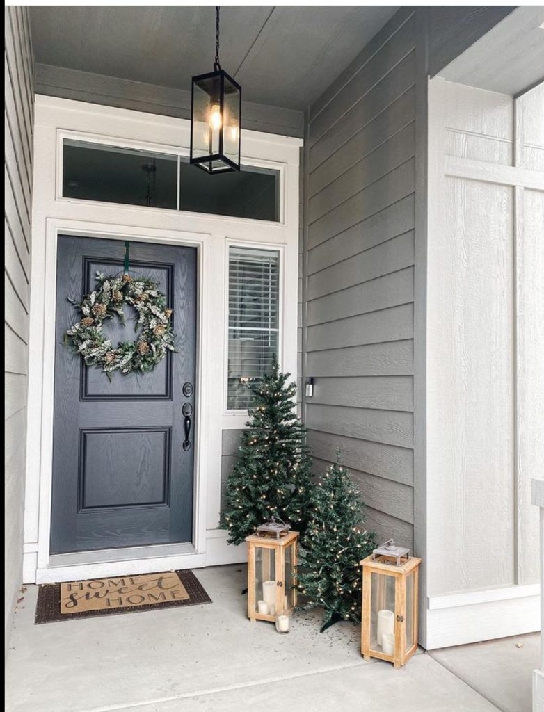 Dark gray front door with a frosted pine cone wreath, flanked by two small pre-lit Christmas trees and two tall light wood lanterns with candles.