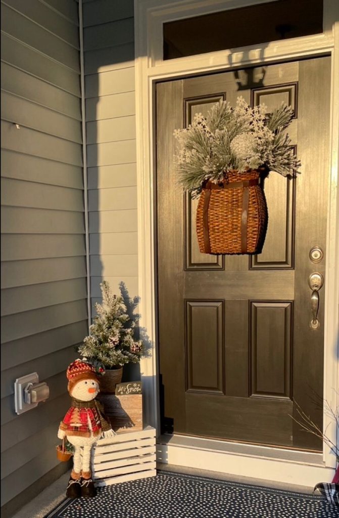 Dark door with a woven basket wreath, a snowman figurine, and a small flocked tree placed on a white wooden crate next to the doorway.