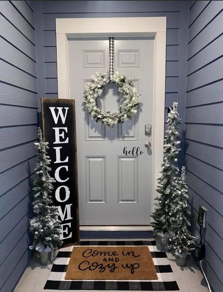 Light gray door with a flocked white wreath, flanked by a tall wooden 'WELCOME' sign and two flocked Christmas trees in galvanized buckets, with a layered doormat.