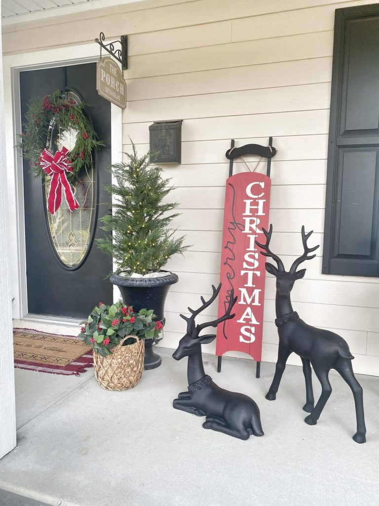 White siding porch with a black door, decorated with a traditional red and green wreath, a large potted tree, a red 'MERRY CHRISTMAS' sign, and two black reindeer figurines.