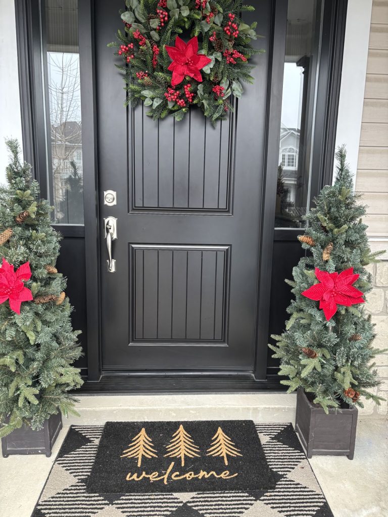 Black front door with a wreath accented with red poinsettias and berries, flanked by two potted Christmas trees similarly accented with red poinsettias and pine cones.