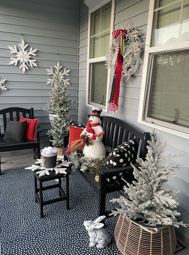 Covered porch with a black bench and polka-dot pillows, a snowman figurine, and several flocked Christmas trees in baskets, with large wooden snowflakes hanging on the wall.