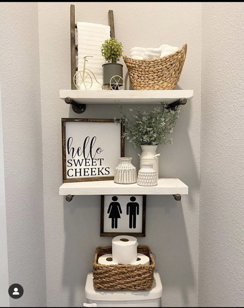 Rustic farmhouse bathroom decor with two pipe-mounted wooden shelves above the toilet, featuring a woven basket, a small ladder, signs, and a bike sculpture.
