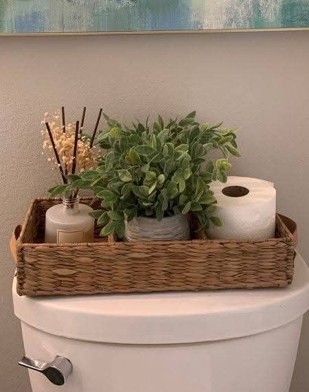 Close-up of a woven natural tray on a white toilet cistern, holding a potted faux plant, a room diffuser, and a roll of toilet paper.