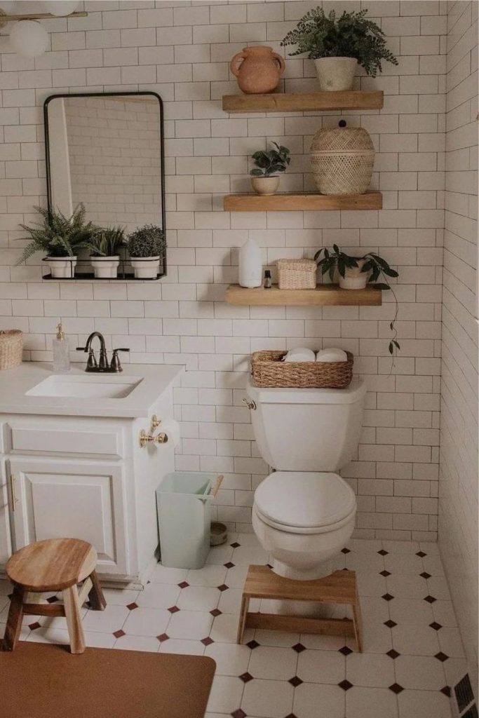 Bright bathroom with subway tiles, three wooden floating shelves filled with potted plants, and a woven basket on the toilet cistern, with a small wooden stool.