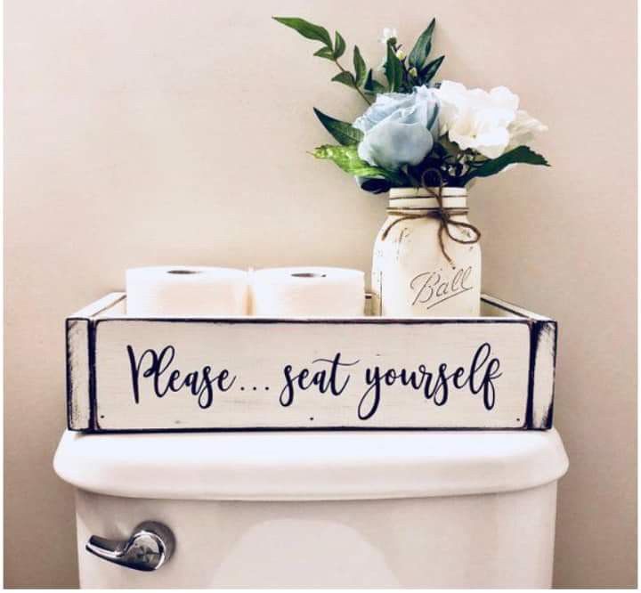 Charming farmhouse toilet cistern decor with a white wooden crate saying "Please... seat yourself," holding toilet paper, and a Mason jar with blue and white flowers.