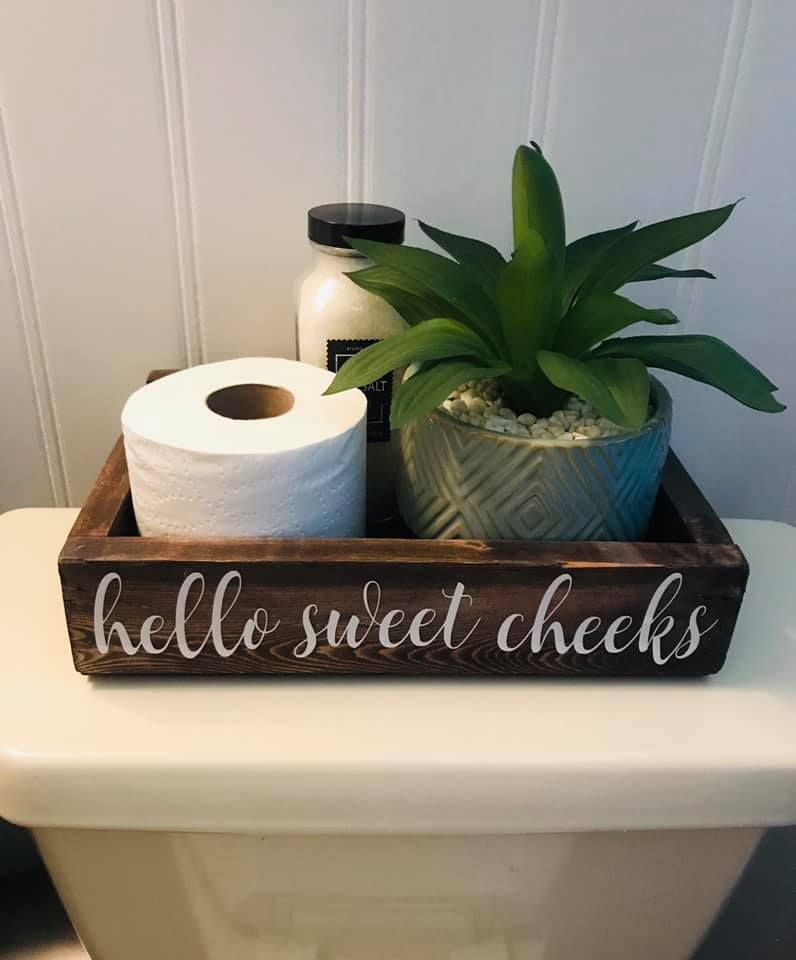 Close-up of a rustic wooden tray on a toilet cistern, inscribed with "hello sweet cheeks," holding a roll of toilet paper, a potted plant, and a bottle of bath salts.