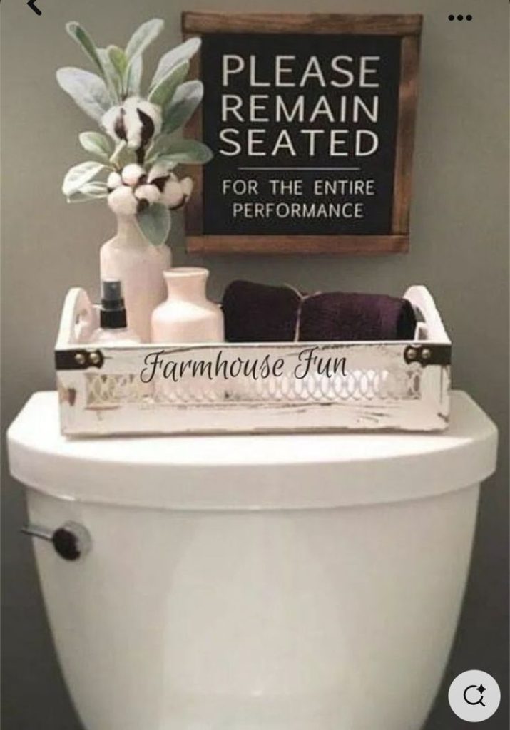 Whitewashed wooden tray on a toilet cistern holding dark folded towels, small vases with faux cotton, and a humorous sign above that reads "Please Remain Seated."