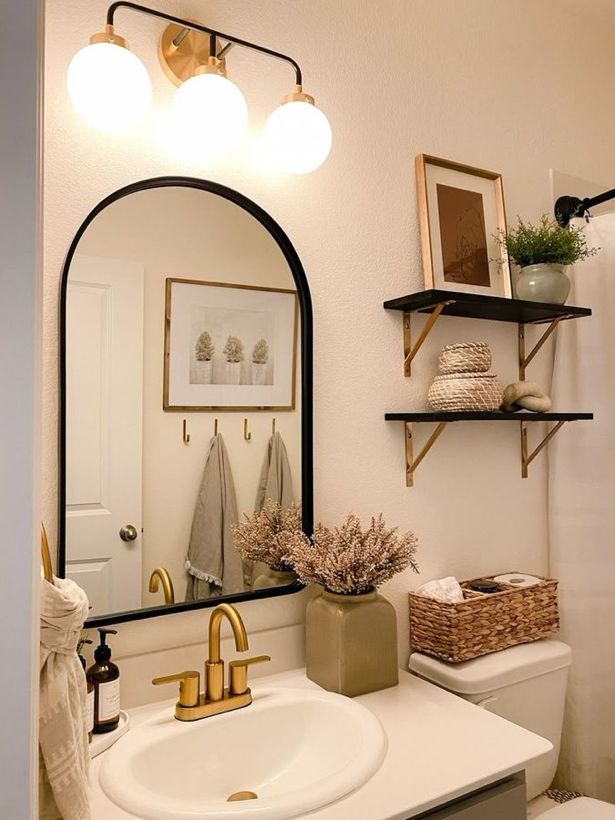 Elegant bathroom decor with brass light fixture and arched mirror, a woven storage box on the toilet cistern, and small stacked baskets on black floating shelves.