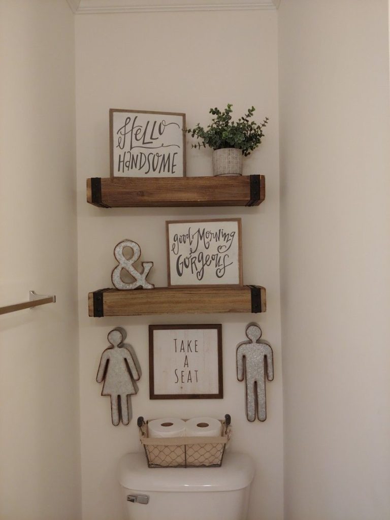 Cozy greetings and dual shelves in a farmhouse bathroom, featuring wooden signs and a wire basket for toilet paper on the cistern.