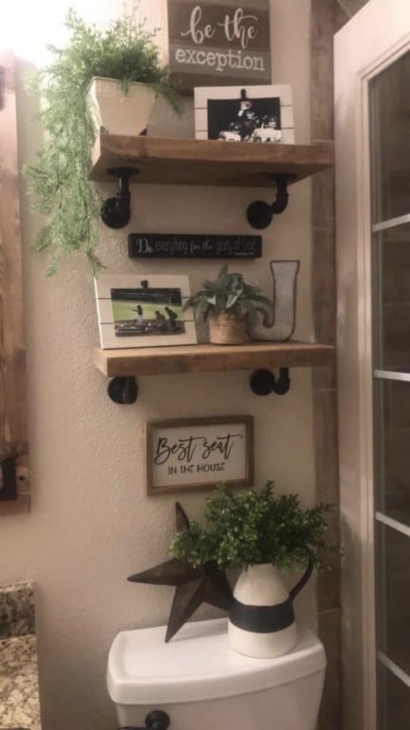 Industrial pipe shelves and rustic charm in a bathroom, with framed photos, signs, and greenery on shelves above the toilet.