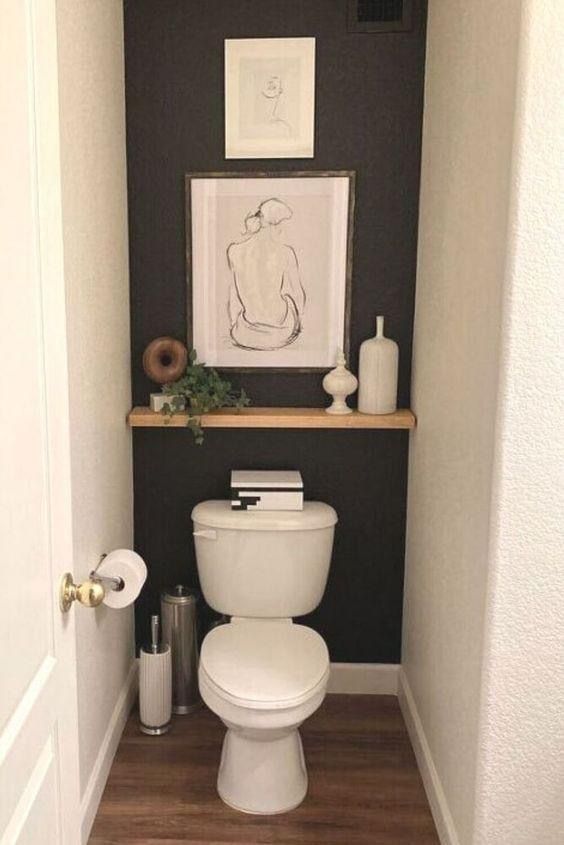 Dramatic black wall and simple art nook in a small water closet, with a wooden shelf and stacked black and white boxes on the toilet cistern.