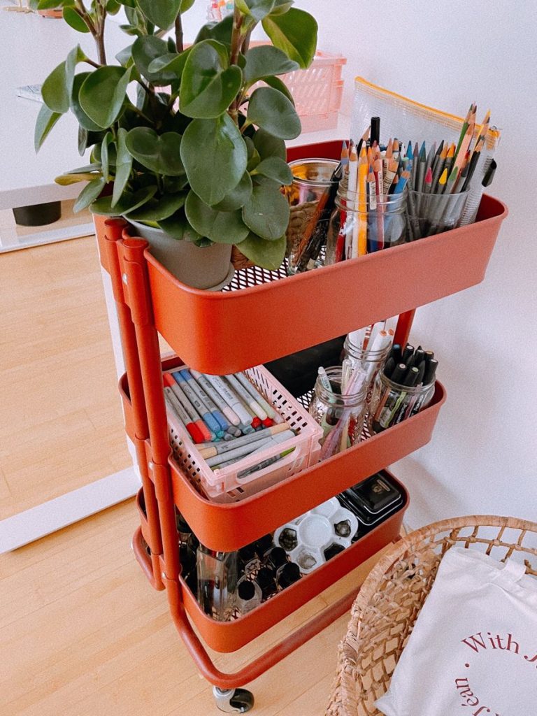 A terracotta-colored three-tier rolling cart holding art supplies, markers, and a green houseplant.