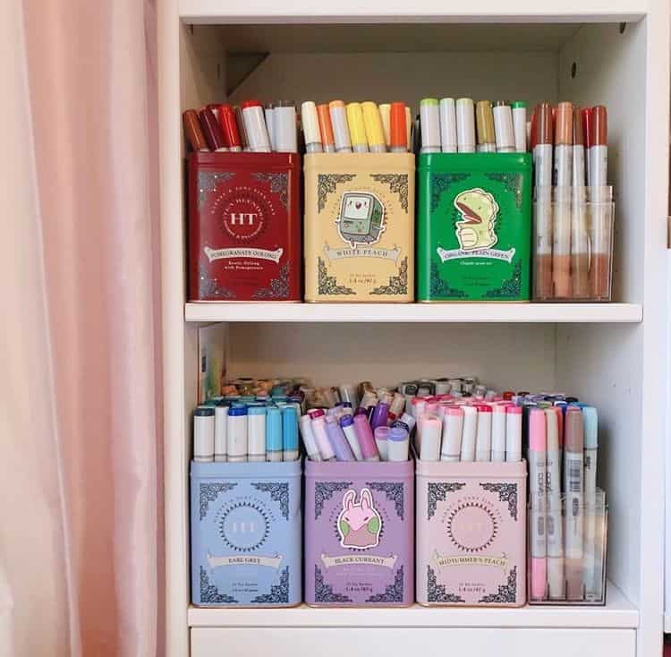 Colorful pastel tea tins filled with markers and pens arranged on a white bookshelf.