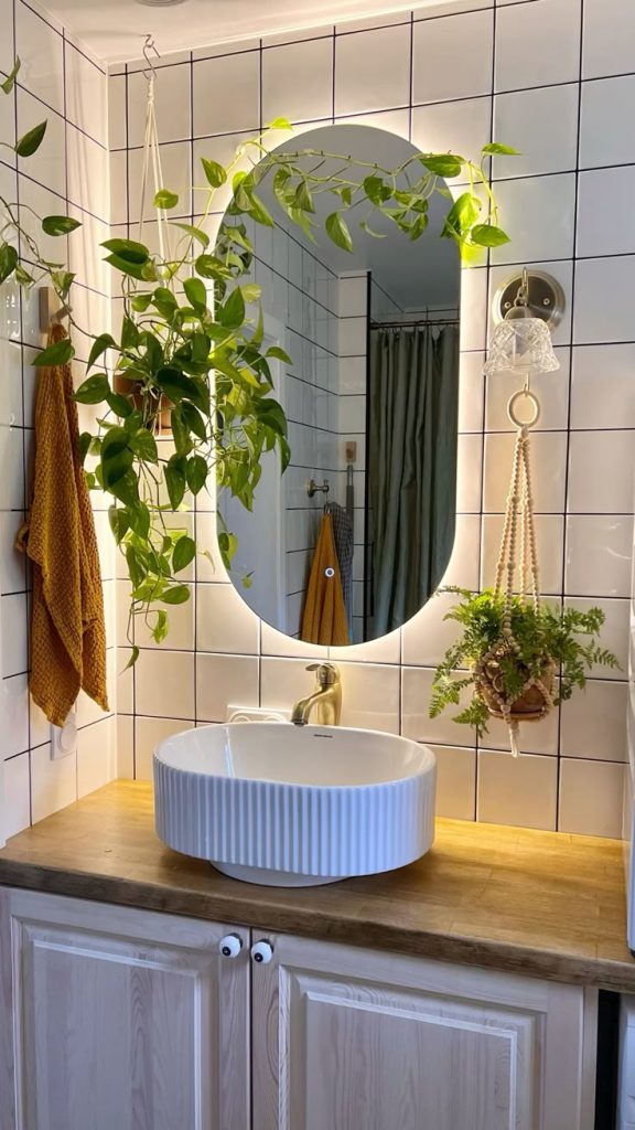 Boho bathroom with a backlit oval mirror, trailing pothos plant in a macrame hanger, a ribbed vessel sink on a wood vanity, and an amber waffle towel.