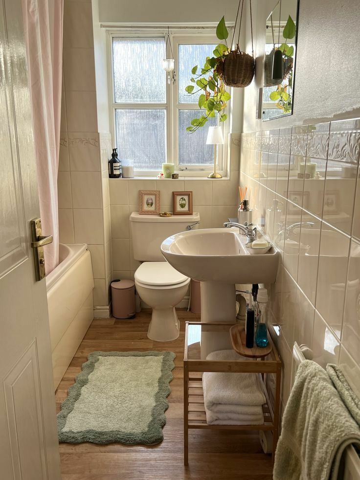 Soft neutral bathroom with small vintage style frames propped on the windowsill, a hanging wicker plant basket from the ceiling, a scalloped edge bath mat, and a small bamboo sink shelf underneath.