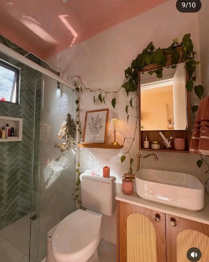 Dreamy boho bathroom with trailing pothos vines draped across the wall, a wooden framed botanical art print on a shelf above the toilet, a rattan medicine cabinet with a mirror, and a small table lamp for warm lighting.